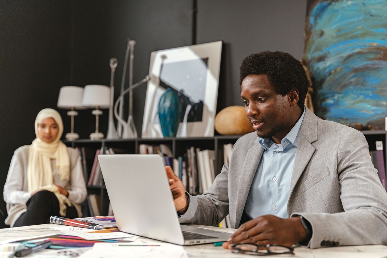 A manager in a grey jacket checks a project on a laptop, a colleague behind him