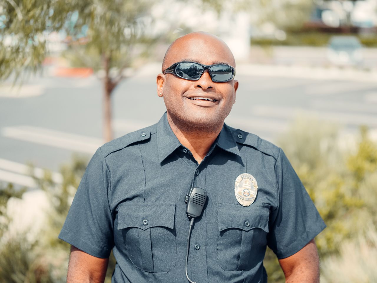 A security officer in uniform, with a radio on his chest, smiles outside a building